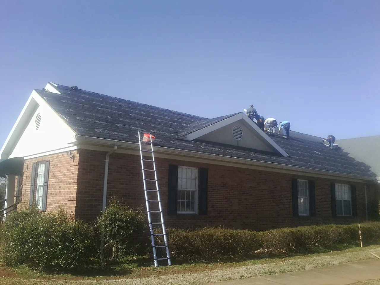 Crew tearing shingles off a brick ranch roof with a ladder set — Metal Roof Repair work by our crew in Wainscott