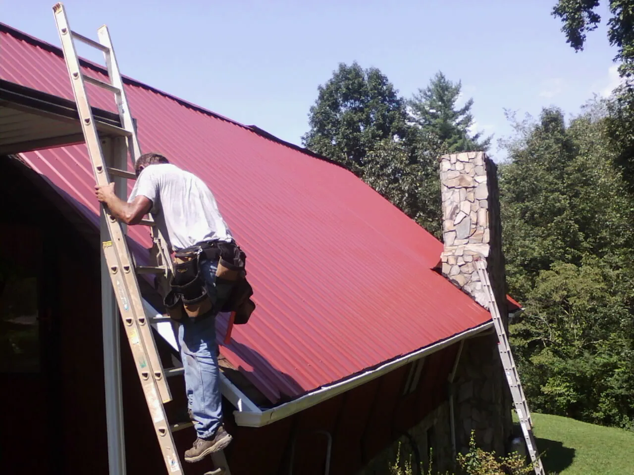 Roofer on a ladder beside a red metal roof and stone chimney — Soffit & Fascia Repair by Roofing Brothers in Wainscott